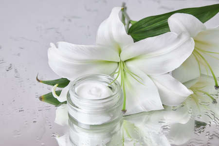 Beauty jar cream cosmetic with lily flowers lie on table, jar cream product on wet flower mirror moistening. Natural flower jar cream cosmetics for hand skin care. Smooth beauty skinの写真素材