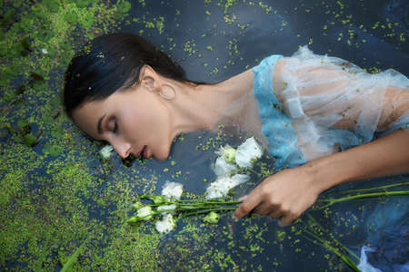 Art beautiful romantic woman lies in swamp in blue long dress with flowers. Portrait brunette in transparent dress in water swamp mud duckweed. Book coverの写真素材