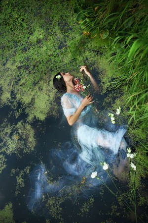 Art beautiful romantic woman lies in swamp in blue long dress with flowers. Portrait brunette in...