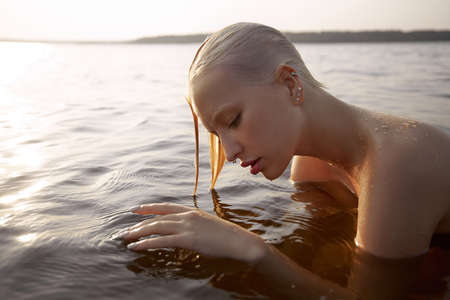 Nude naked sexy woman in water at sunset. Beautiful blonde woman with short wet hair and big breasts, art portrait in seaの写真素材