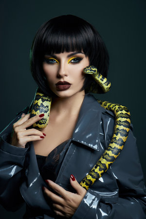 Beauty woman python yellow snake around her neck on latex shiny raincoat. Yellow snake on the shoulders of girl. Beauty yellow eye shadow makeup, dark burgundy lipstickの写真素材