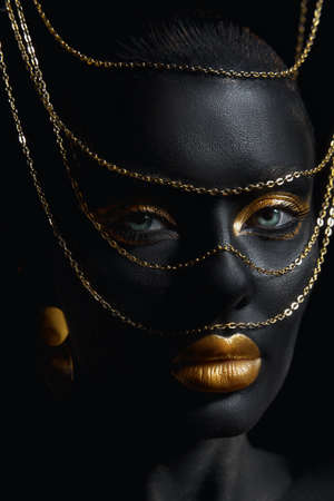 Beauty woman painted in black skin color body art, gold chain in his hands and around his neck. Gold...