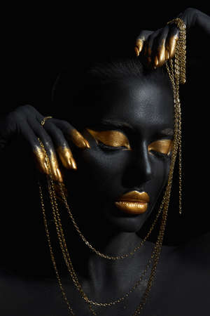 Beauty woman painted in black skin color body art, gold chain in his hands and around his neck. Gold makeup lips eyelids, fingertips nails in gold color paint. professional makeupの写真素材