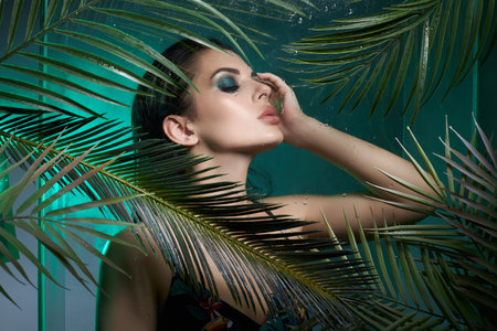 Beauty tropical portrait of a woman in palm leaves, wet long hair, green eye shadow makeup