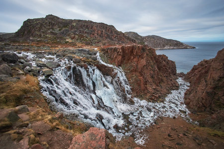 Waterfall tumbles over rocks into the ocean under a cloudy sky as evening approaches. Teriberka, Russiaの写真素材