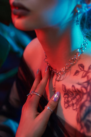 Woman with blue and red lighting on her face poses in front of green foliage. She is wearing multiple pieces of jewelry and has a tattoo on her chestの写真素材