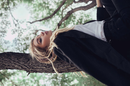 Blonde woman in dreadlocks leans against tree in woods, wearing blazer, t-shirt, and pants with eyes closed and mouth slightly open.の写真素材