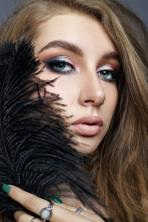 Beauty woman with long brown hair and striking blue eyes. Woman is wearing colorful makeup and a black feather is partially covering her faceの写真素材