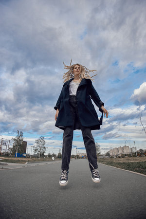 A young woman wearing a dark coat and black pants jumps in the air with her blonde hair blowing in...