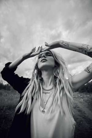A woman with long blonde dreadlocks and several necklaces stands in a field, looking up at the cloudy sky. Her hands are raised, shielding her eyes from the sun.の写真素材