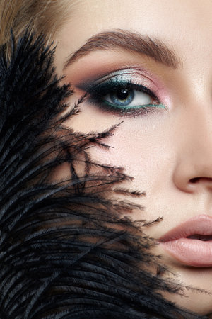Beauty woman with long brown hair and striking blue eyes. Woman is wearing colorful makeup and a black feather is partially covering her faceの写真素材
