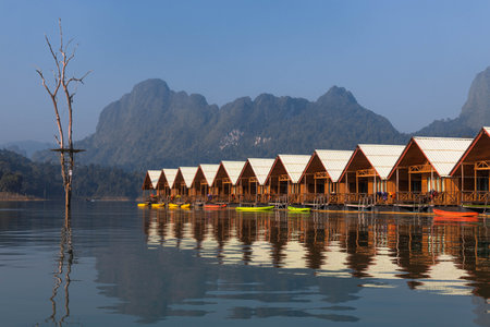 Wooden cabins sit on still waters, reflecting the surrounding mountains and trees. Kayaks are lined up along the shore, inviting exploration of this serene natural setting.の写真素材