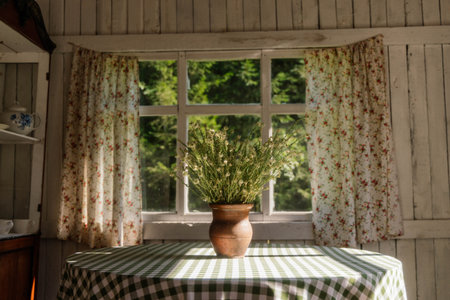 Sunlight filters through floral curtains, brightening a cozy kitchen featuring a round table with a checkered cloth and a vase filled with vibrant herbs, inviting warmth and homey charm.の写真素材