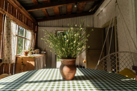 A beautifully arranged clay vase holds fresh flowers, placed on a green and white checkered table in a rustic interior, bathed in warm daylight.の写真素材
