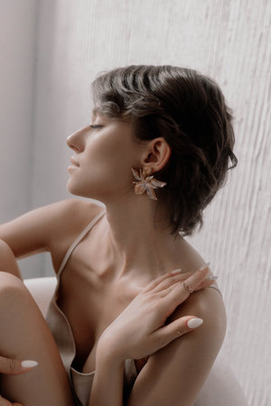 Beauty woman with a chic short haircut showcases beautiful floral earrings while seated elegantly. Soft natural light highlights her serene expression, enhancing the overall atmosphere of tranquility.の写真素材