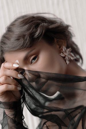 Beauty woman playfully obscures her face with translucent fabric while highlighting her intricately designed floral earrings, set against a gentle, neutral backdrop.の写真素材
