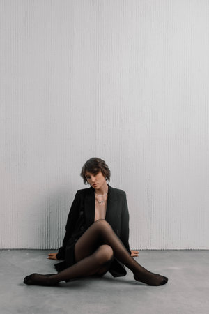 Woman sits gracefully on the floor, wearing a sleek black outfit and hosiery, exuding confidence and style against a simple, textured wall.の写真素材
