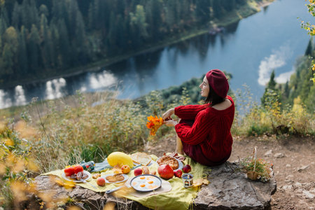 A woman in a cozy red sweater sits by a tranquil lake, surrounded by vibrant autumn foliage while enjoying a picnic laid out on a blanket with various fruits and treats.の写真素材