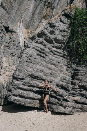 A woman stands elegantly beside majestic rock formations at a tranquil beach, basking in the midday sun. The lush greenery and smooth sand enhance the natural beauty of the moment.の写真素材