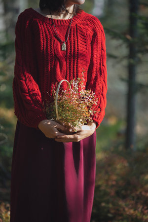 A woman stands amidst tall trees in a forest, wearing a cozy red sweater. She cradle a basket filled with wildflowers, enjoying the peacefulness of the golden hour.の写真素材