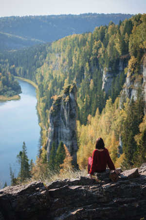 Contemplative woman sits on a rock ledge, gazing out over a serene river cutting through a vibrant, forested landscape filled with autumn colors.の写真素材