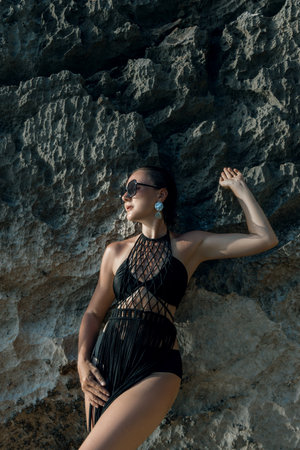 A woman wearing a stylish black swimsuit leans against a rugged rock formation at the beach, showing her confidence under the warm sun, enjoying a carefree summer moment.の写真素材