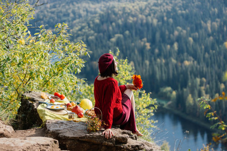 A woman sits on a rocky ledge, admiring the picturesque view of a calm lake framed by vibrant autumn trees. A picnic spread rests on the ground nearby, enhancing the peaceful atmosphere.の写真素材