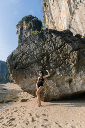 A woman stands confidently next to a large rock formation on a tranquil beach, enjoying the sunshine and natural beauty of her surroundings. The ocean gently laps at the shore.の写真素材