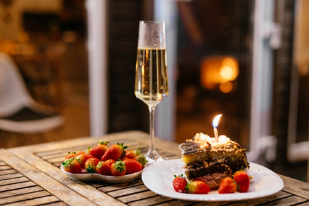 gourmet cake adorned with a candle rests on a plate beside a bowl of fresh strawberries, paired with a sparkling drink, all set on a rustic wooden table outdoors in the evening.の写真素材