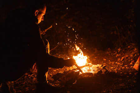 A person kneels beside a glowing campfire, carefully arranging sticks to maintain the flames.