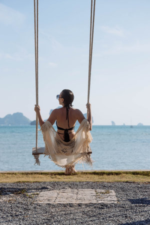 woman sits on a swing, facing the calm sea. She wears a bikini and a light cover-up, soaking in the sun and admiring the distant islands under a clear blue sky.の写真素材