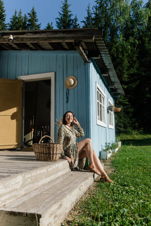 A woman sits on the porch of a charming blue cabin, basking in the sun on a warm afternoon. Surrounded by trees, she holds a basket, seemingly relaxed and content in nature.の写真素材