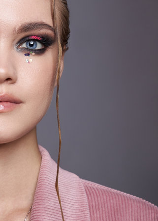 model highlights her artistic makeup with shimmering eyeshadow and gemstones under soft lighting, showcasing a modern fashion aesthetic against a neutral background.の写真素材