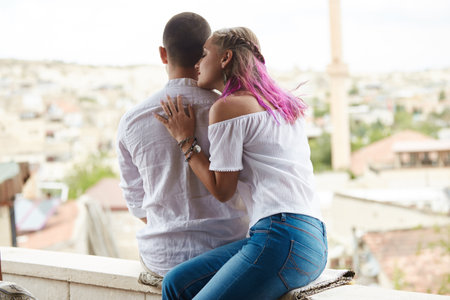 Young couple shares a tender moment while sitting on a ledge, with her pink-haired head resting gently on his shoulder. They overlook a vibrant city landscape under a partly cloudy sky.の写真素材