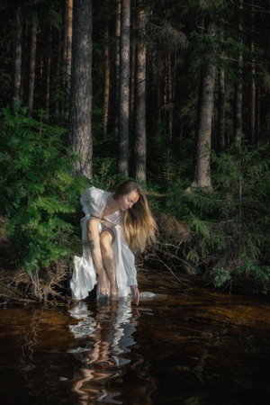 woman with long hair kneels by the edge of a serene water body, gently touching the surface. Tall trees surround the area, casting shadows as the sun begins to set.の写真素材