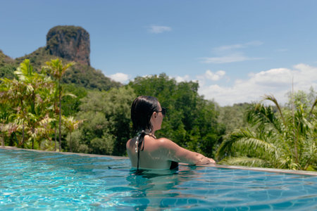 woman enjoys a serene moment in a freshwater pool, surrounded by lush greenery and a stunning mountain landscape under a clear blue sky.の写真素材
