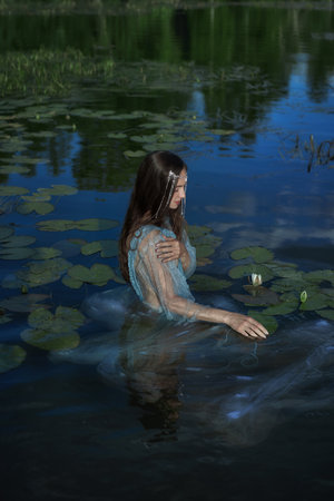 woman dressed in a delicate gown stands gracefully in a calm pond, surrounded by lily pads, reflecting the soft light of the setting sun.の写真素材