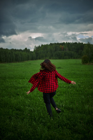 young person in a red plaid jacket spins playfully in a vibrant green meadow, surrounded by trees, as dark clouds gather overhead, creating a serene, adventurous atmosphere.の写真素材