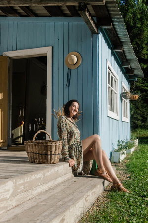 A young woman relaxes on the steps of a charming blue cottage, smiling while basking in sunlight on a warm day, with a wicker basket beside her.の写真素材
