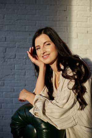 woman with long dark hair is sitting against a white brick wall, smiling gently while resting her...
