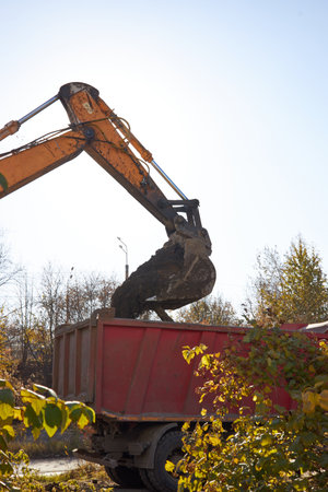 excavator with a large bucket is lifting soil into a red dump truck at a construction site during the day. Clear blue sky is visible above the work area, and trees surround the site.の写真素材