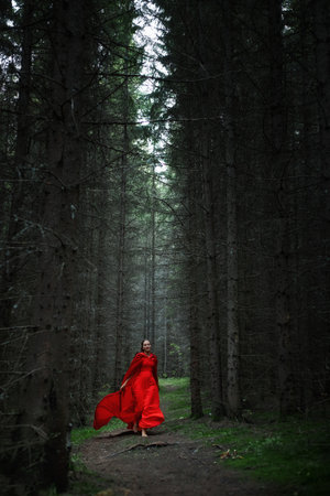 woman in a vibrant red dress glides along a narrow path in a dark, dense forest. Tall trees rise on either side, creating an enchanting and mysterious atmosphere. Soft mist lingers in the air.の写真素材