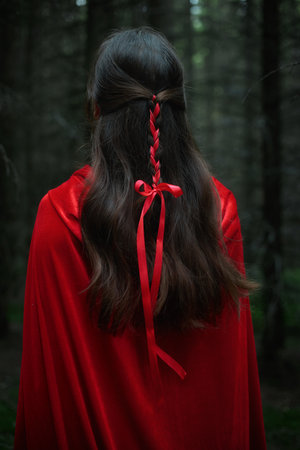 figure in a flowing red cloak stands in a dense forest, facing away from the viewer. hair is styled delicately with a red ribbon, surrounded by tall trees shrouded in soft light.の写真素材