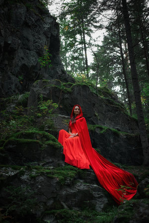 woman dressed in a vibrant red cape sits quietly on a mossy rock in a lush forest. Towering trees and soft light create a magical atmosphere as she gazes into the distance.の写真素材