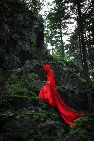 woman wearing a long red cloak gracefully walks down a rocky path surrounded by green moss and trees in a dimly lit forest. atmosphere is serene and mysterious.の写真素材
