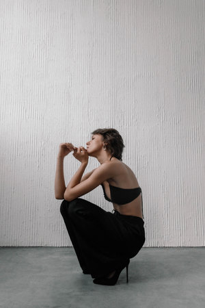 female model is squatting gracefully, deep in thought. She wears a black outfit that showcases her figure, with a textured white wall providing a minimalist backdrop for the scene.の写真素材