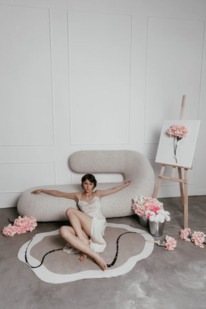 model sits elegantly on a unique, soft couch in a light-filled room, showcasing a delicate outfit while surrounded by floral arrangements and an easel with artwork.の写真素材