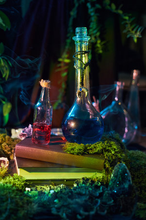 Two glass bottles, one filled with red liquid and the other with blue, rest on moss-covered books. environment is dark, with hints of greenery and smoke, enhancing a magical atmosphere.の写真素材