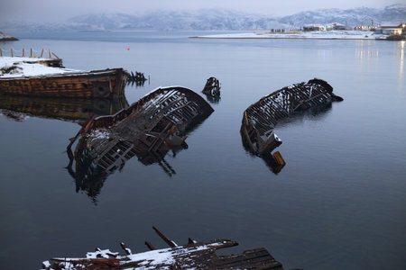 In Teriberka, Russia, old wooden boats lie wrecked in shallow water. Snow covers the landscape, and...