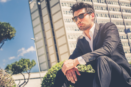 Handsome man model sitting with sunglasses. Career and job opportunities. Young entrepreneur, behind him a building with offices.の写真素材